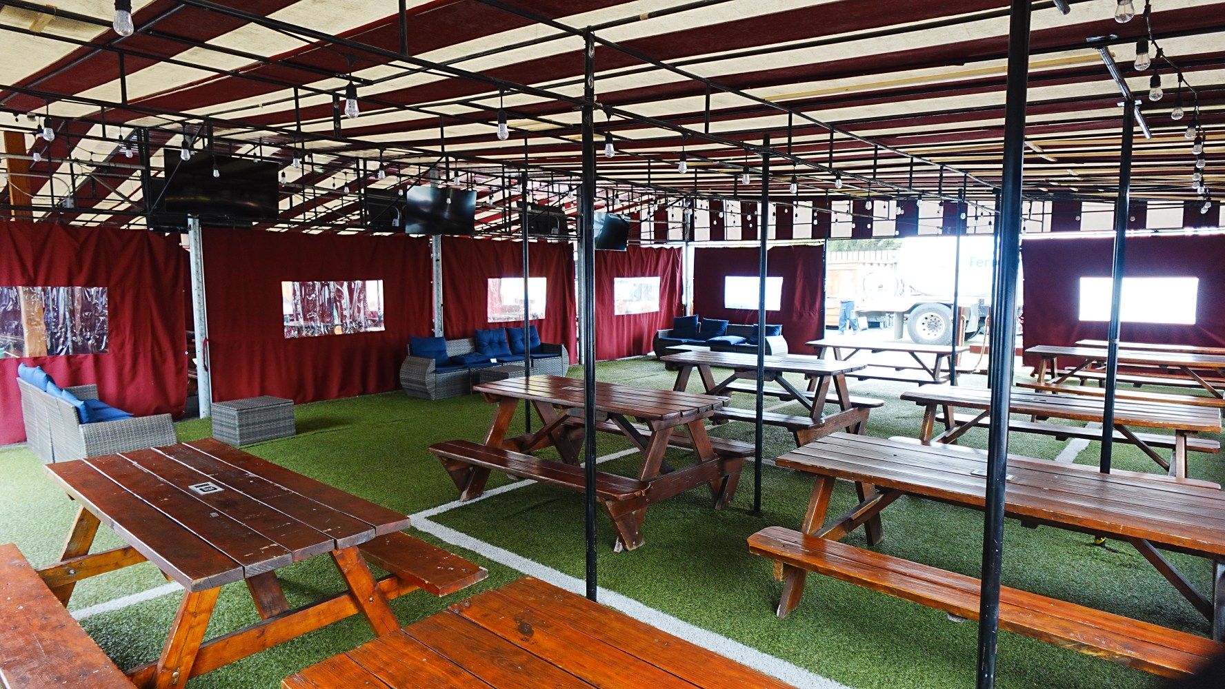 Outdoor seating area with wooden tables, benches, red curtains, and artificial grass floor.