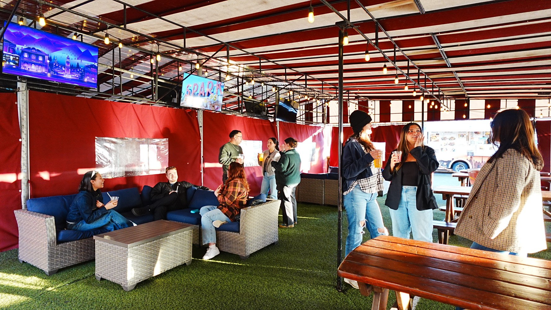 People chatting in an outdoor covered area with seating and a TV.