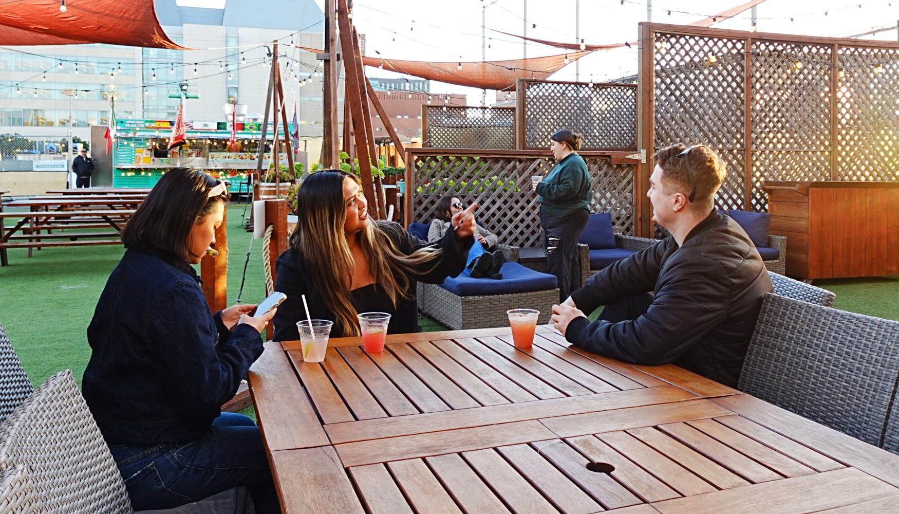 Three people sitting at an outdoor wooden table with drinks, enjoying a sunny afternoon.