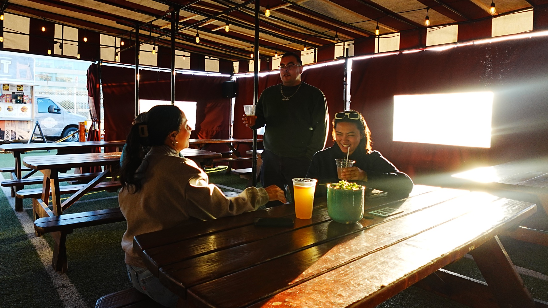 Three people at a picnic table with drinks in a sunlit patio.