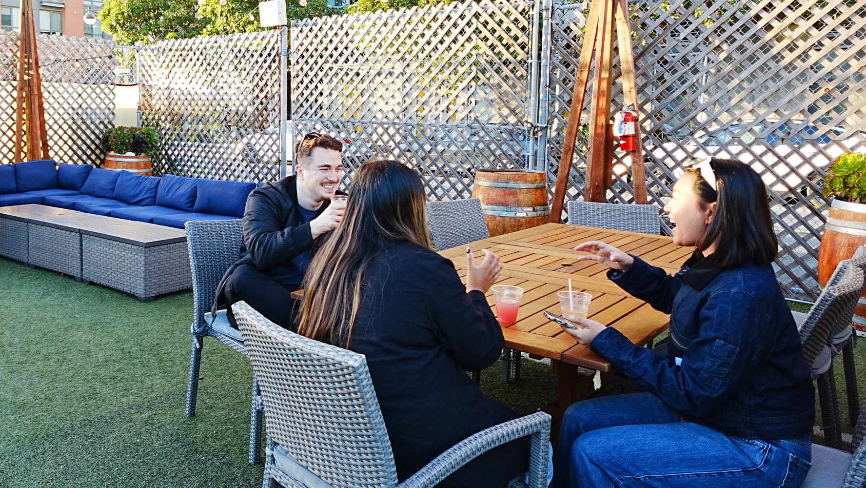 Three people laughing around a table outdoors with drinks.