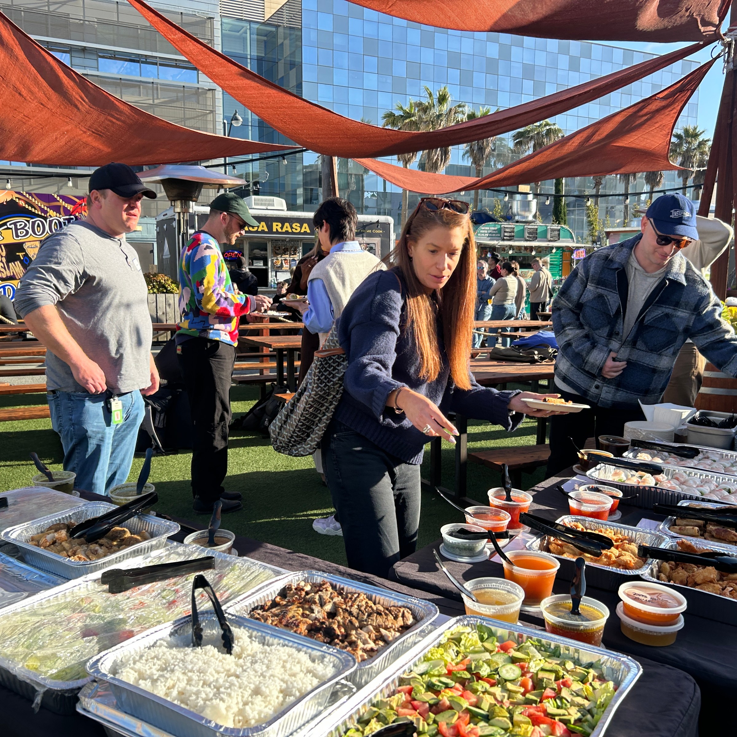People serving food from a buffet outdoors with orange canopies overhead.