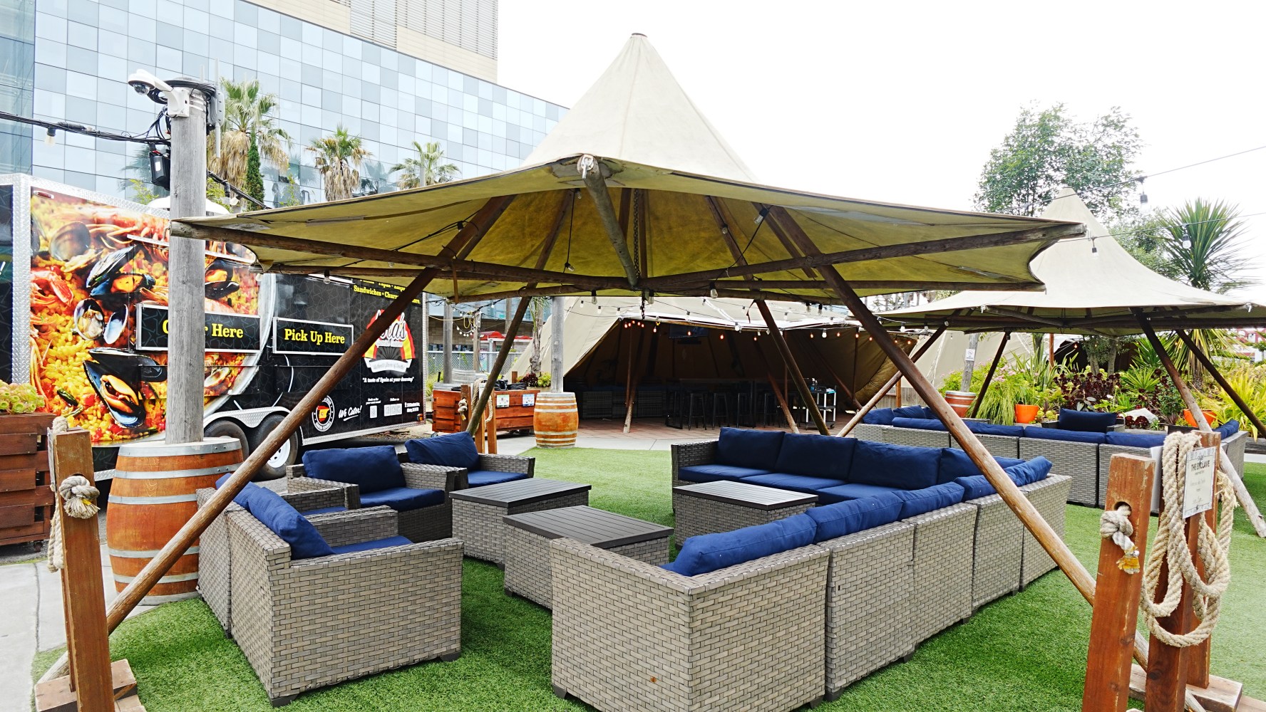 Outdoor seating area with shaded tents, wicker sofas, and a palm tree in the background.