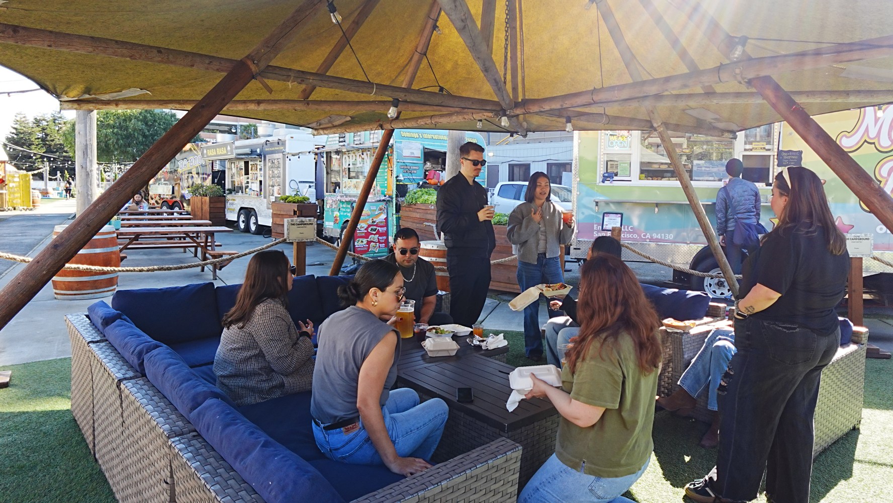 People seated and standing under a canopy in an outdoor lounge area.