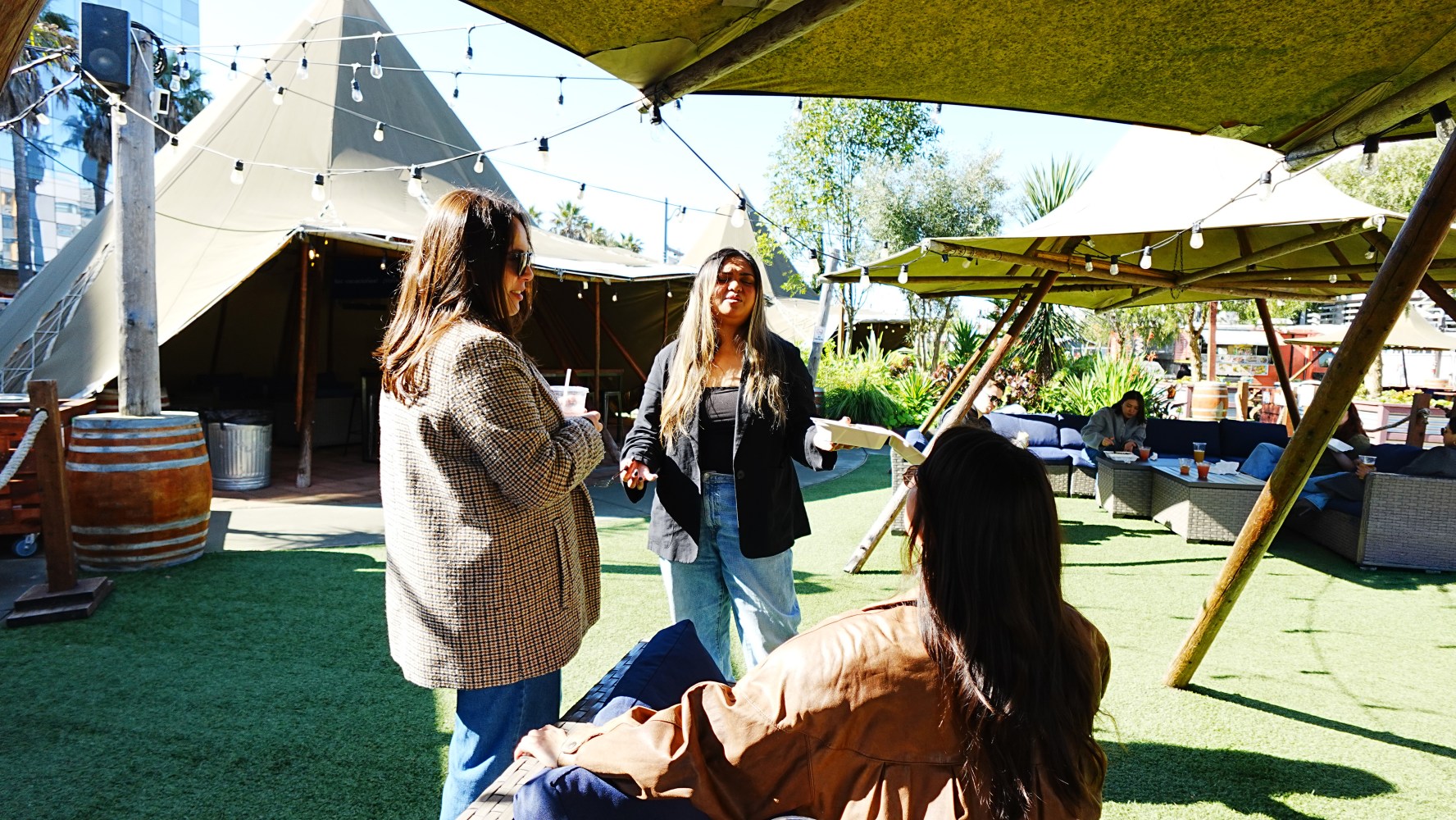Three people talking under a tent with string lights in a sunny outdoor area.