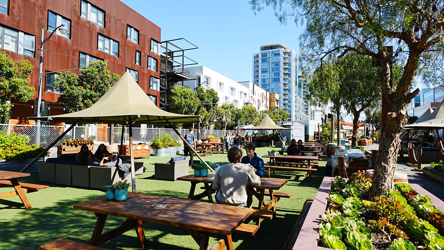 Urban park with benches, umbrellas, trees, and people on a sunny day.