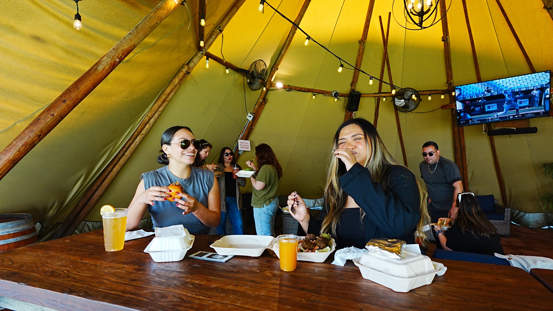 Two women enjoy food and drinks at a wooden table inside a tent with hanging lights.