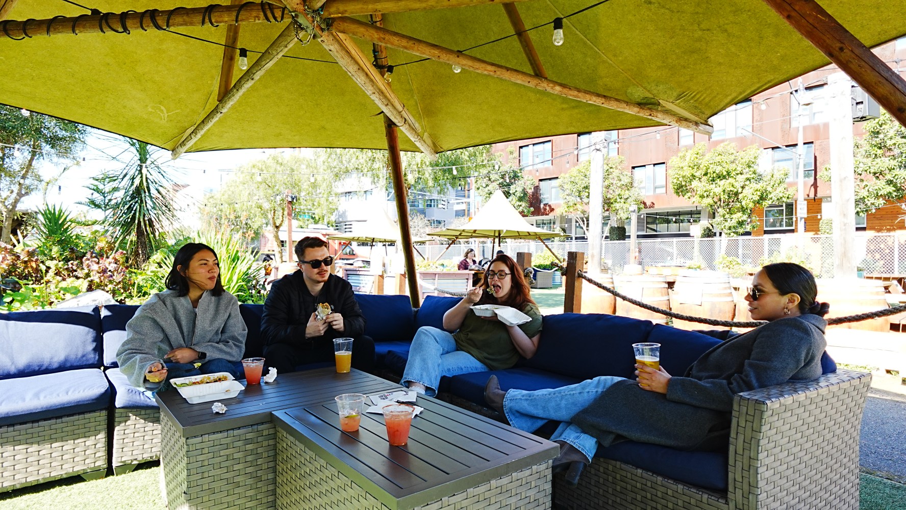 Four people relaxing under a large umbrella on outdoor couches with drinks.