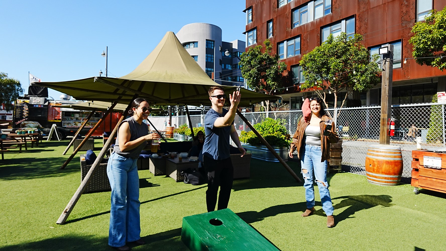 Three people play cornhole outdoors near a building and large tent.