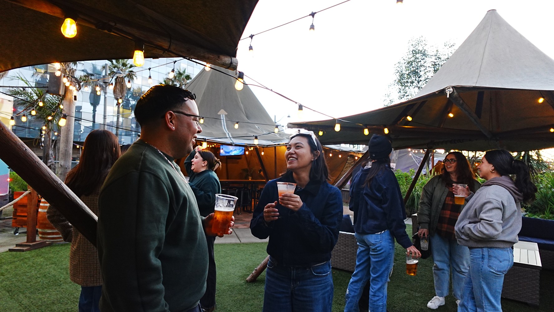 People socializing at an outdoor venue with string lights and tents.