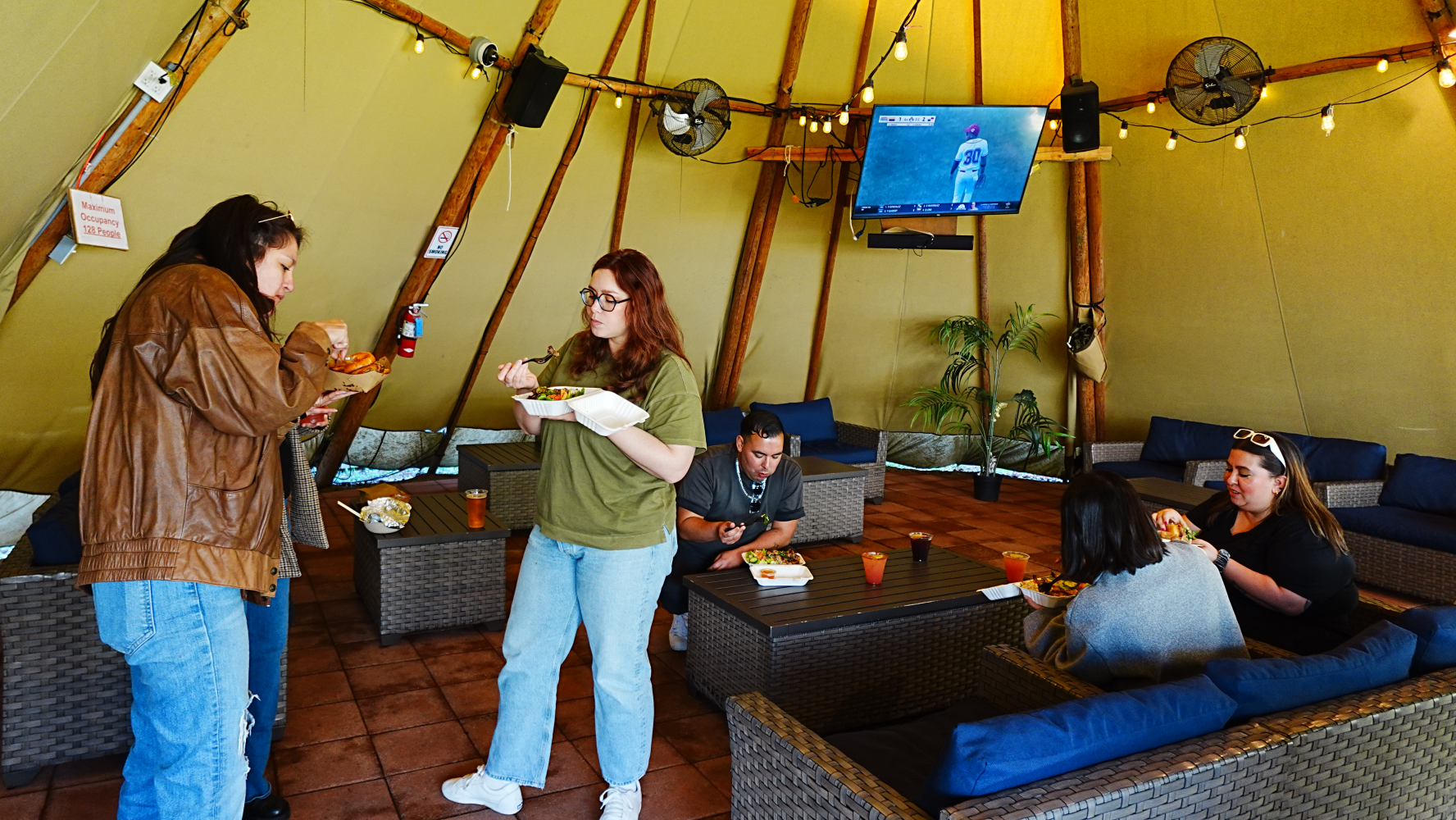 People eating and socializing inside a tent with a TV on the wall.