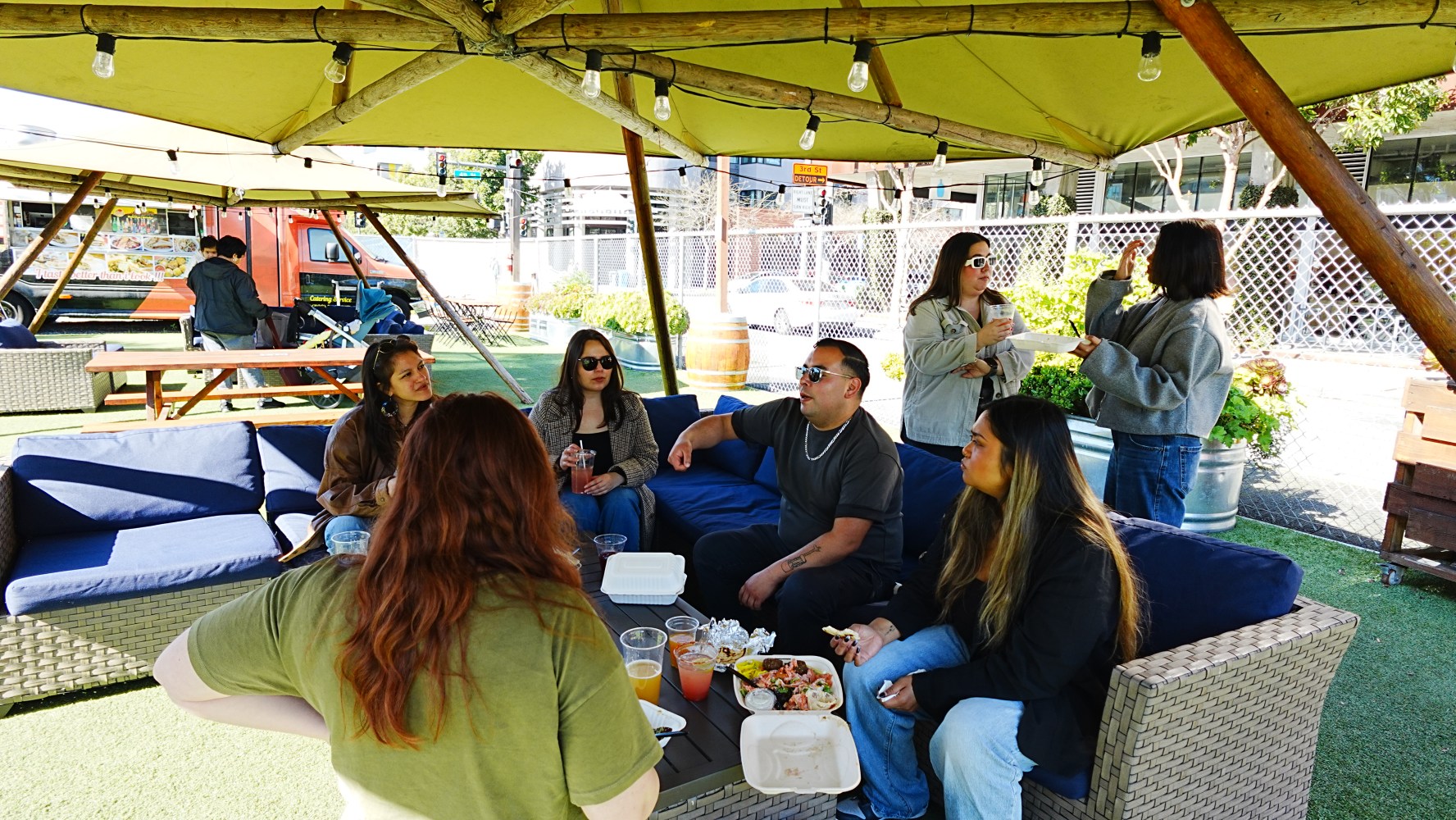 Group of people sitting on outdoor sofas under a gazebo, eating and talking.