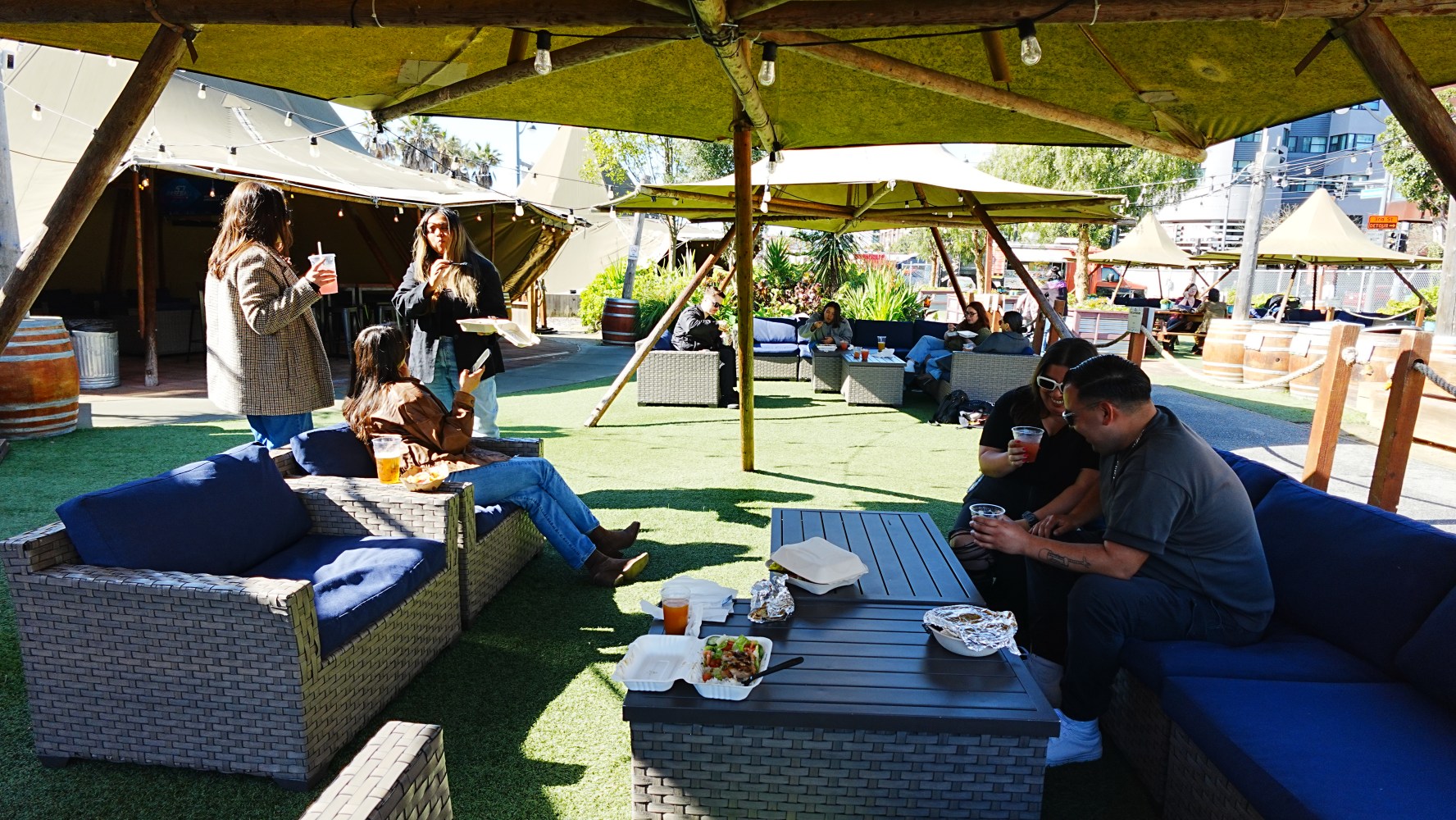 People relaxing on outdoor sofas under tents in a sunny area with food and drinks.