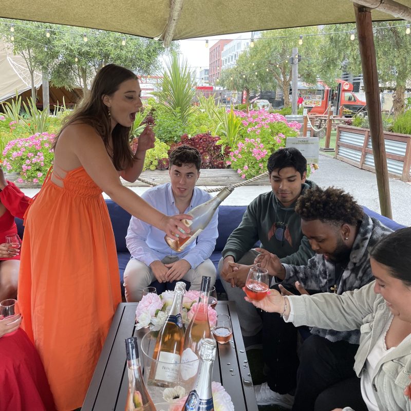 Group of people sitting outdoors with drinks under a tent, surrounded by flowers and greenery.