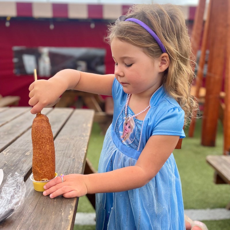 Girl in blue dress holding corn dog over a wooden table outdoors.
