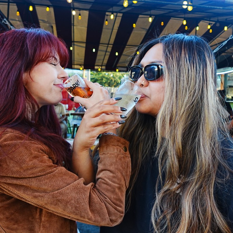 Two women smiling and drinking from glasses in an outdoor setting.