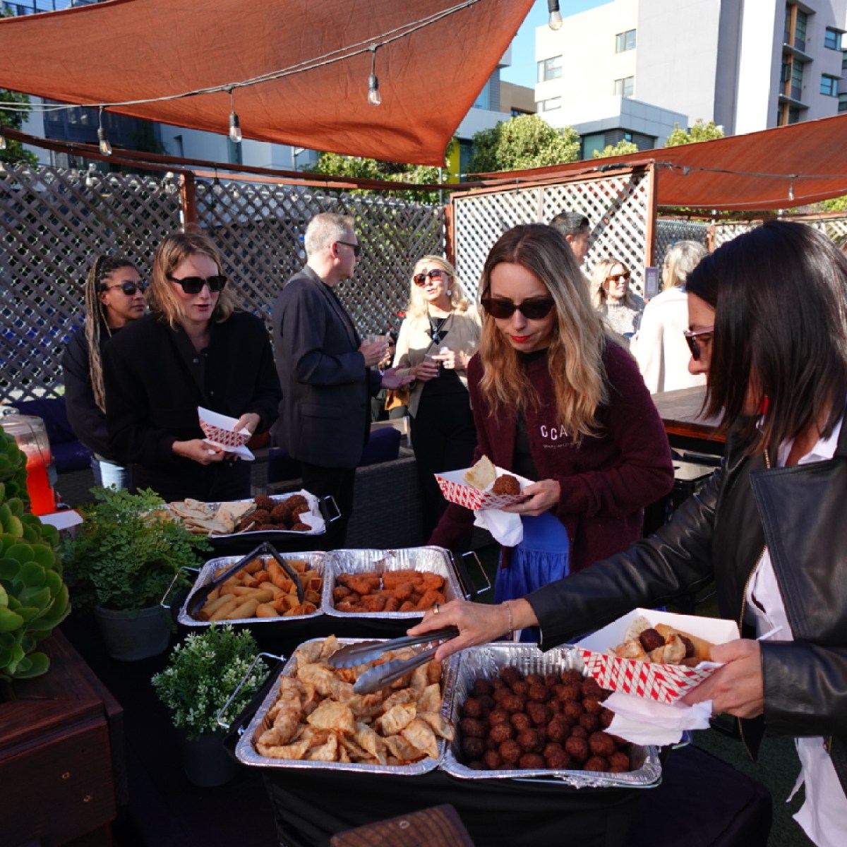People at an outdoor gathering serving themselves food from trays under shade canopies.
