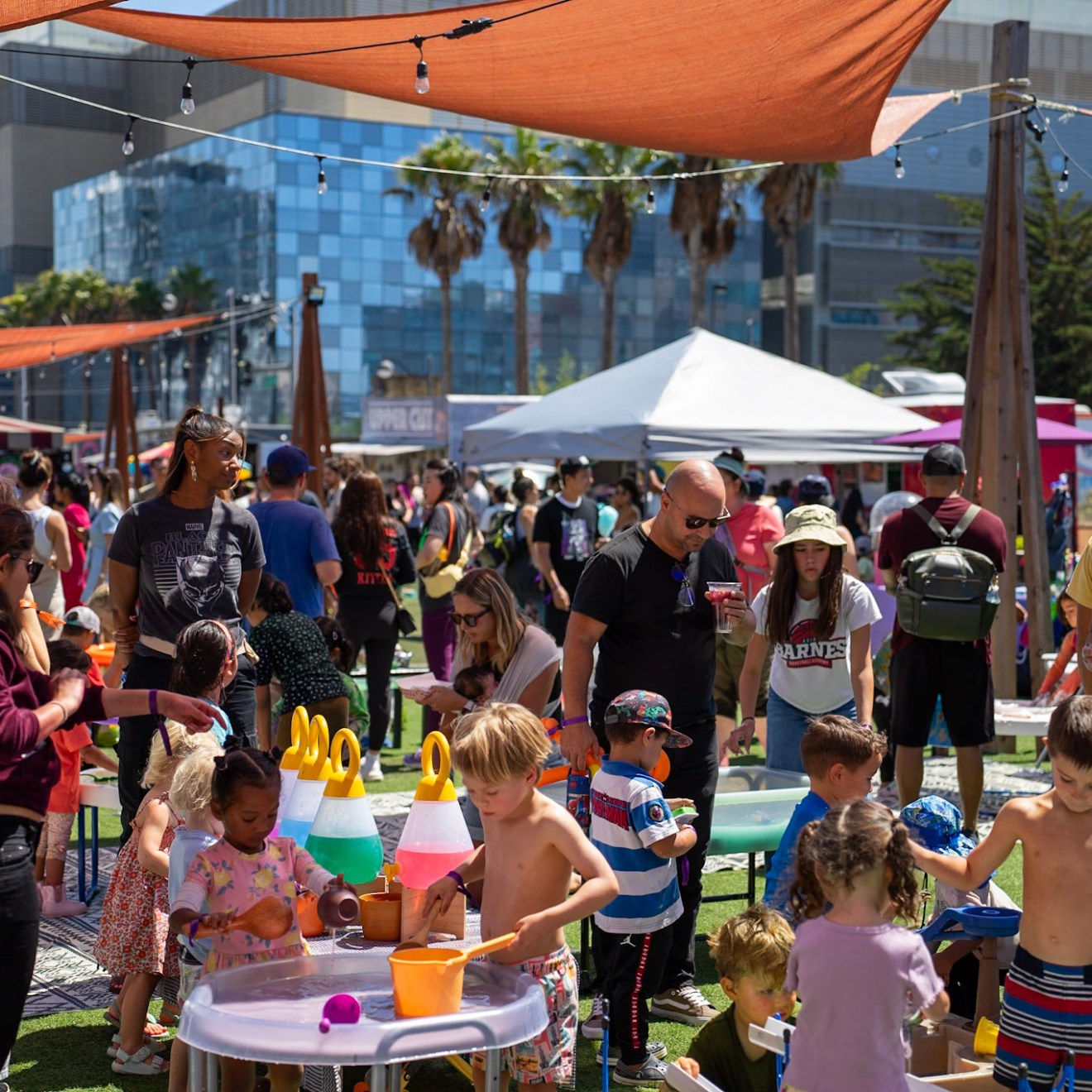 People at an outdoor event with children playing at activity tables, under tents and string lights.