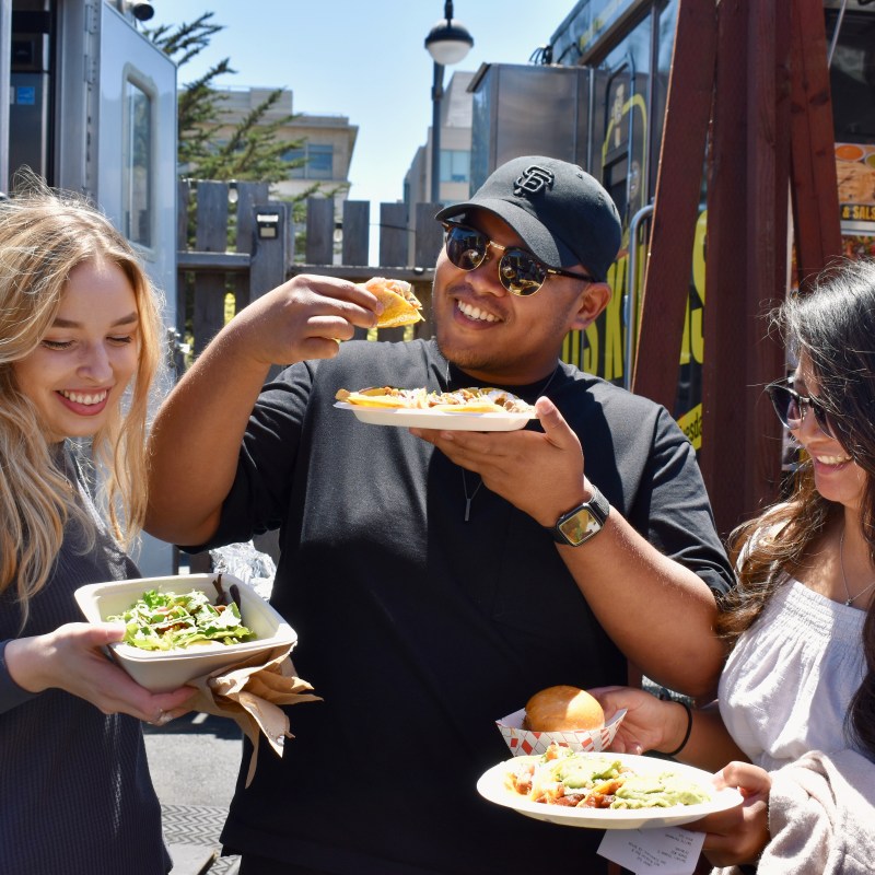 Three friends enjoying food from a food truck, smiling and holding plates with tacos and salads.