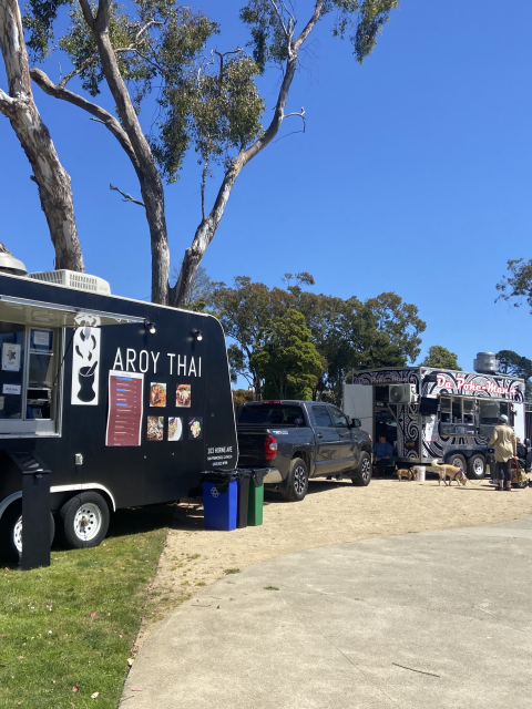 Food trucks parked on a sunny day near trees and tents on a sidewalk.