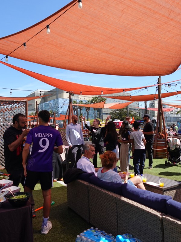 Outdoor gathering with people under orange canopies, food table, and string lights.