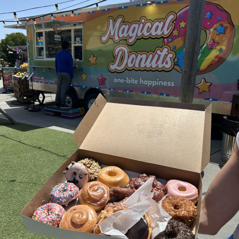 Box of assorted donuts held in front of a colorful Magical Donuts truck on a sunny day.