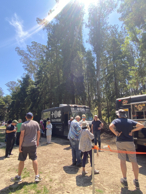 People gather around food trucks in a sunny, wooded park setting.