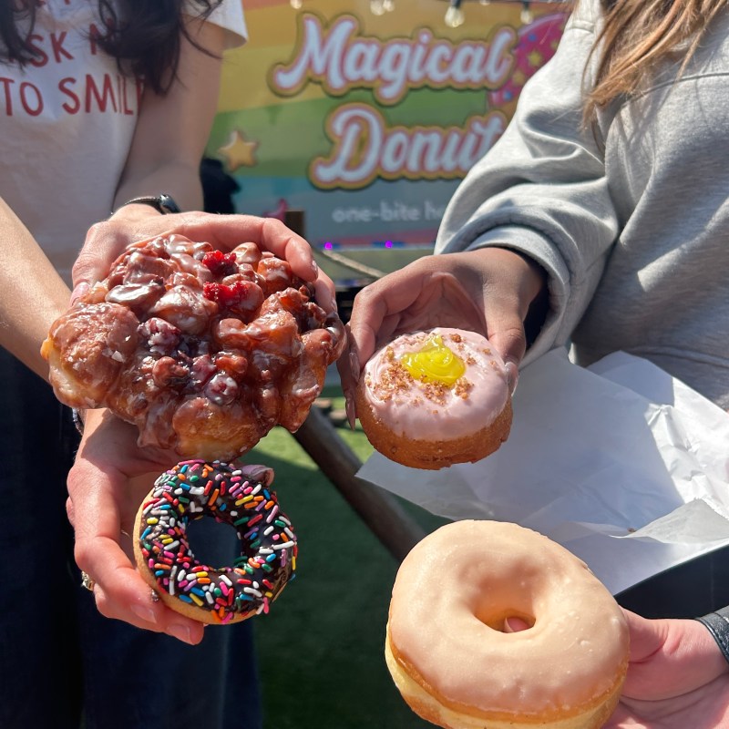 Four people holding different donuts with a colorful 'Magical Donuts' sign in the background.