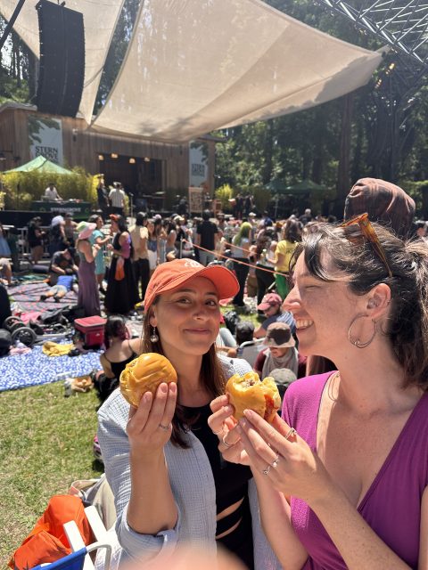 Two people smiling, holding burgers at an outdoor music festival with a crowd and stage in the background.