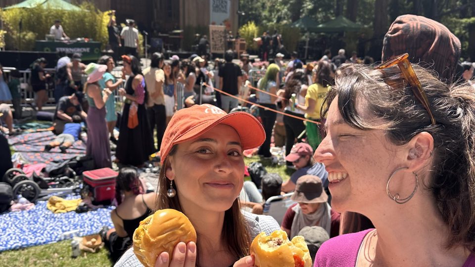 Two people smiling, holding burgers at an outdoor music festival with a crowd and stage in the background.