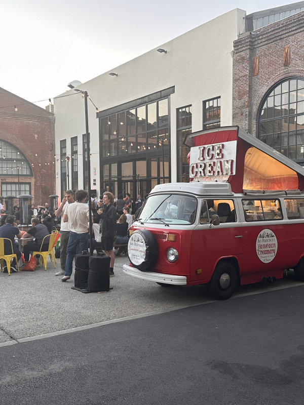 Outdoor gathering with people near a red ice cream van in a courtyard with brick buildings.