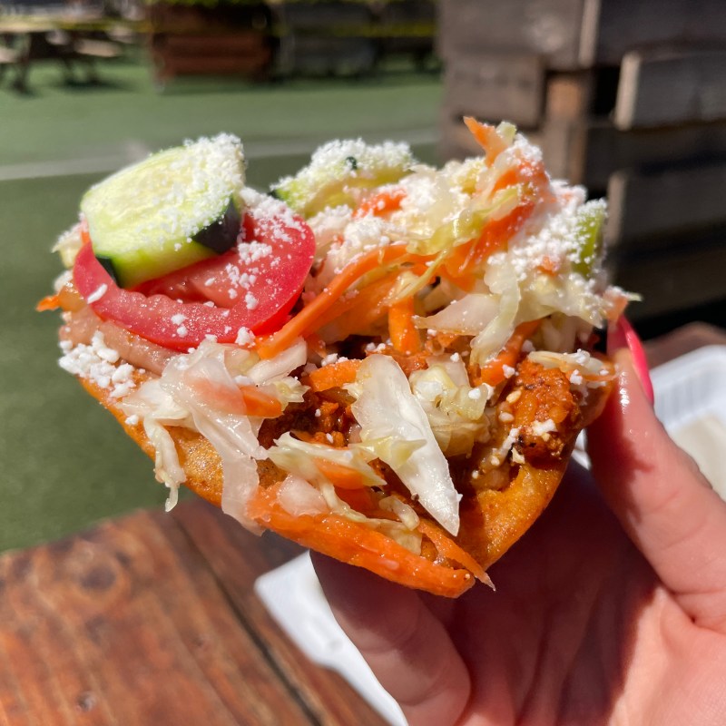 Hand holding a taco with vegetables, cheese, and meat outside on a sunny day.