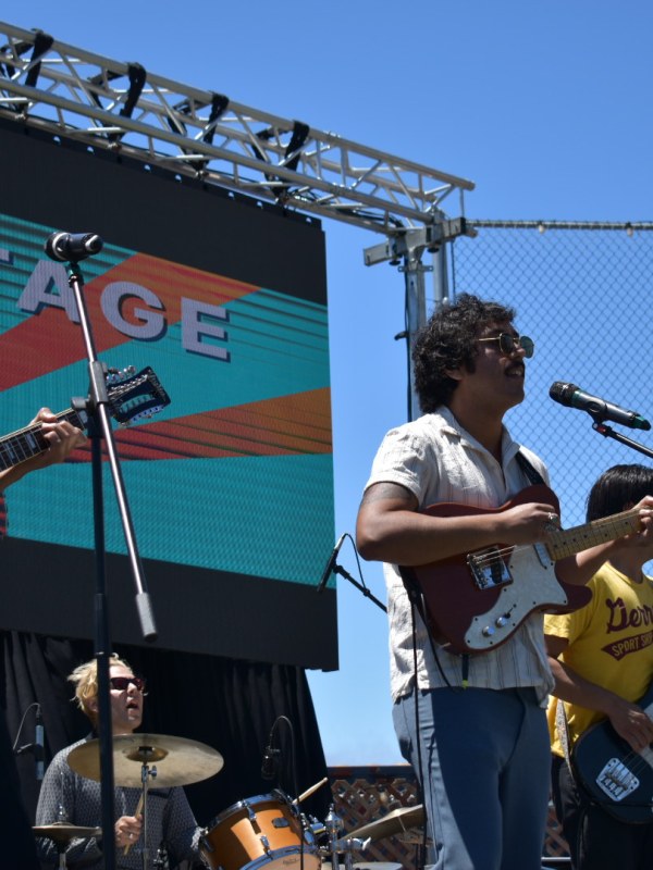 Band performing on outdoor stage with guitars and drums, sunny day.