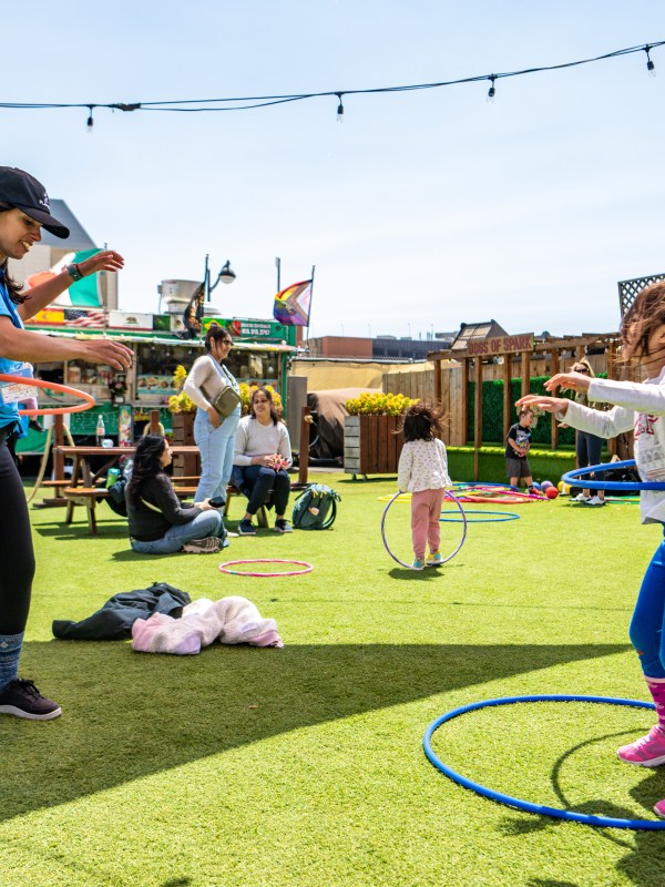 People hula hooping on a sunny, grassy rooftop with string lights overhead.