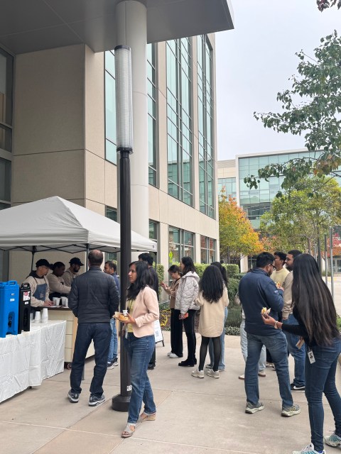 People gathered at a food stall under a white canopy outside a modern office building.