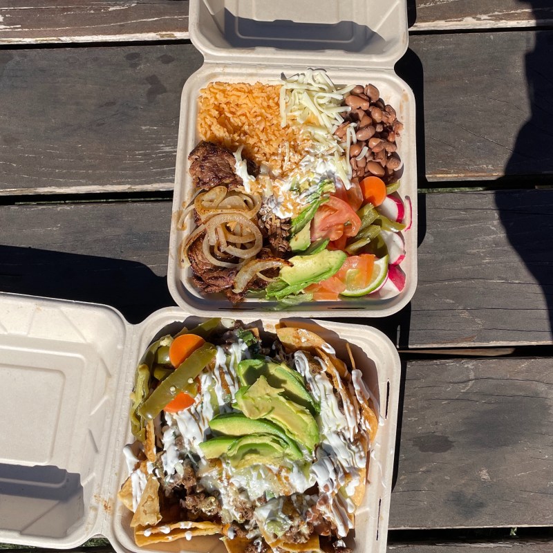 Two takeout boxes: one with rice, beans, veggies, and meat; the other with nachos and avocado on a wooden table.