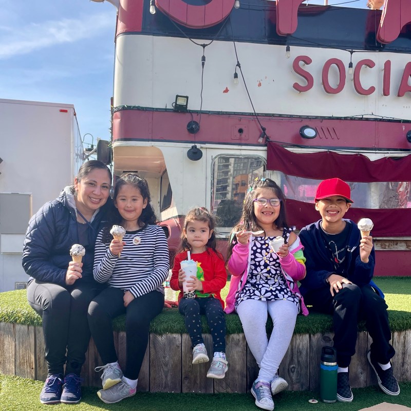 Five people sitting on grass, holding ice creams, outside a red and white building.