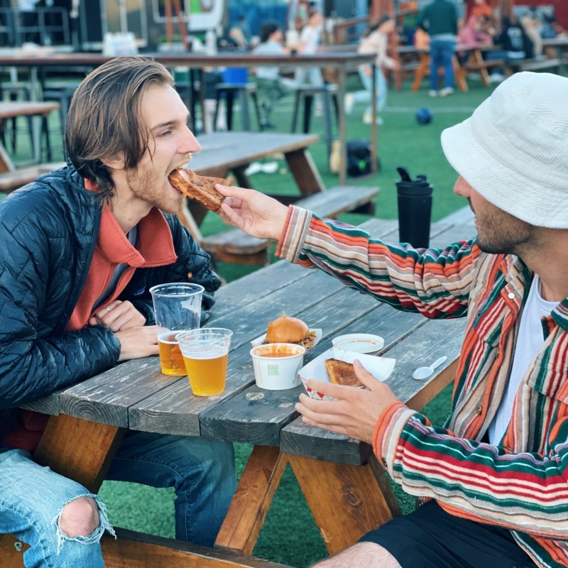 Two people at an outdoor table; one feeds the other a sandwich.