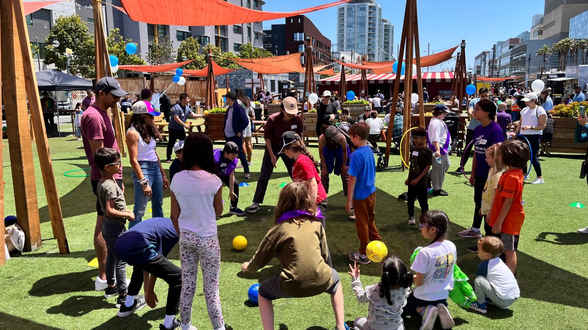 Children playing with a ball in a sunny outdoor area with red canopies and city buildings in the background.