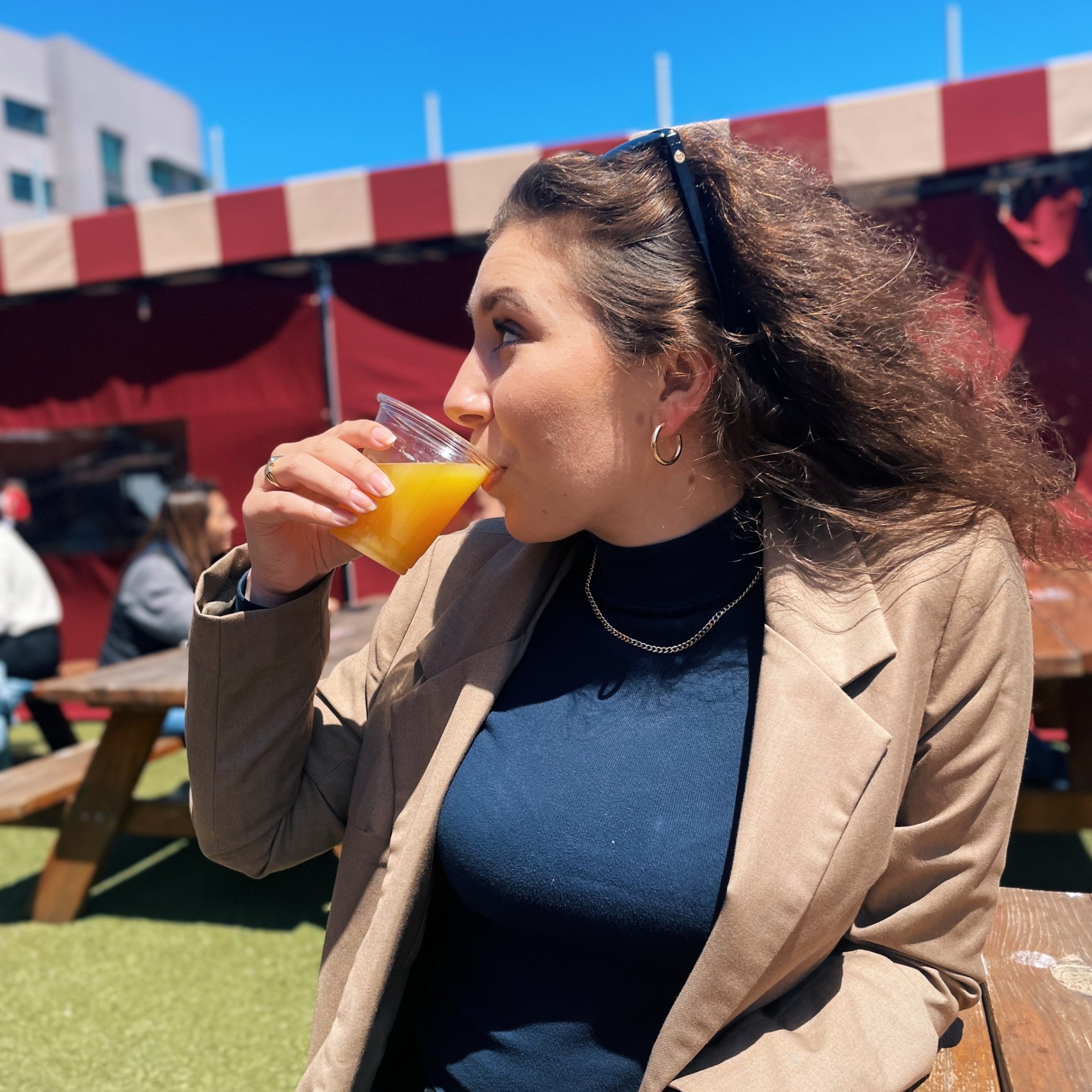 Woman in beige blazer drinking orange juice outdoors on a sunny day.