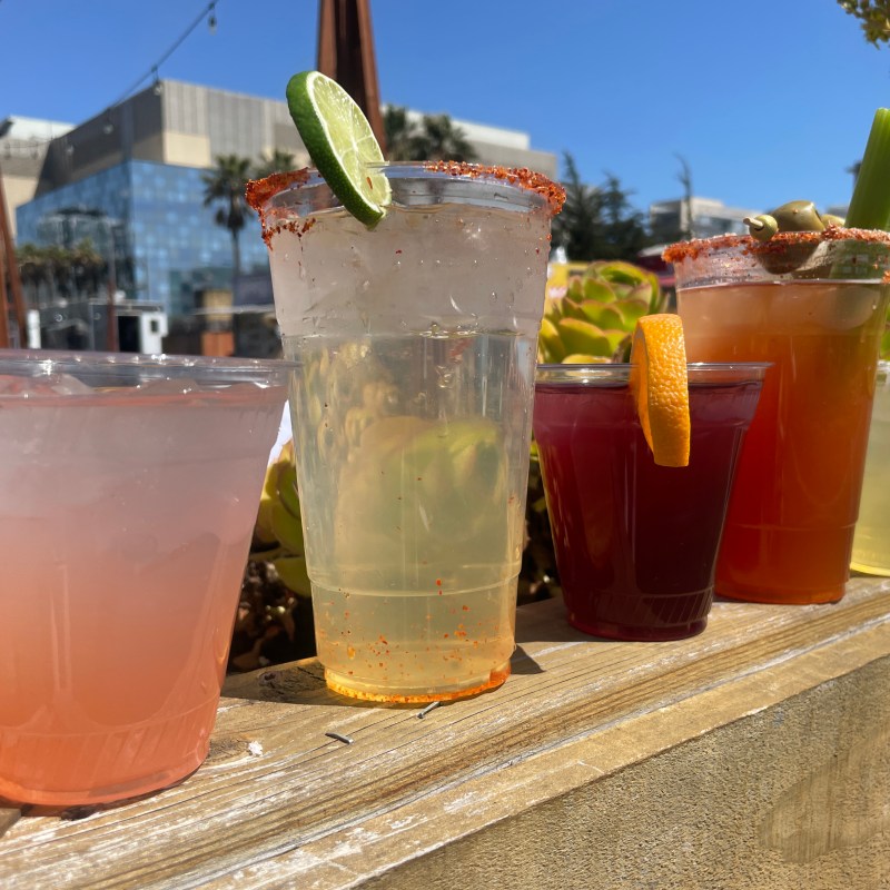 Five diverse cocktails in plastic cups on a wooden surface, with outdoor background.