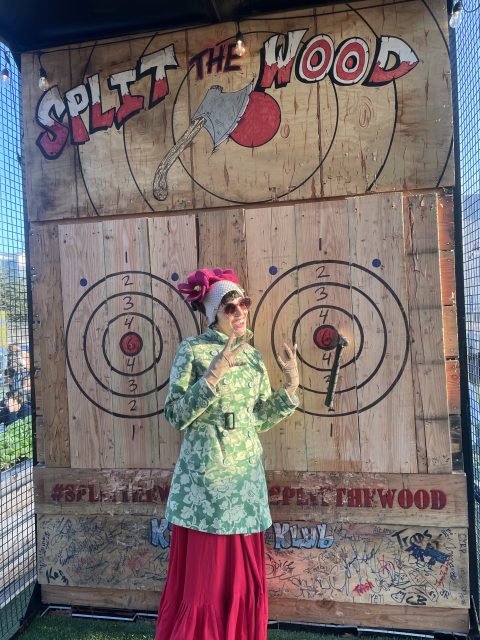 Person in colorful clothing poses near axe-throwing targets labeled 'Split the Wood' with axes in bullseyes.
