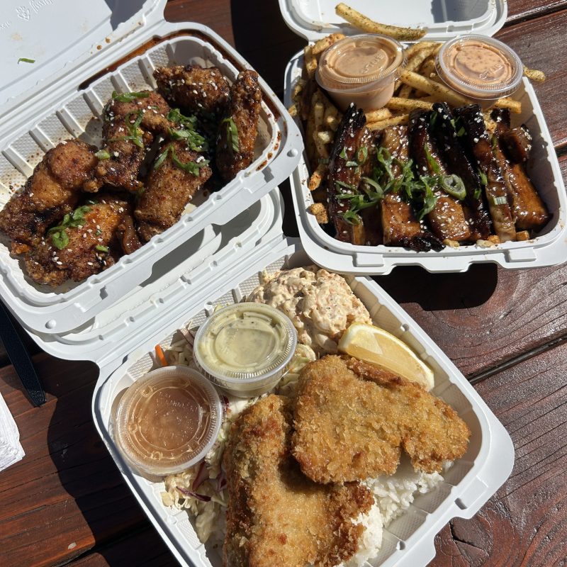 Three takeout trays with fried chicken, glazed ribs, fries, coleslaw, rice, sauces, and lemon wedge on a table.