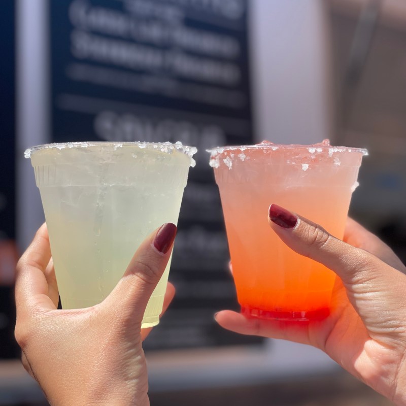 Two hands clinking plastic cups with salted rims, one with yellow liquid, one orange, against a blurred menu.