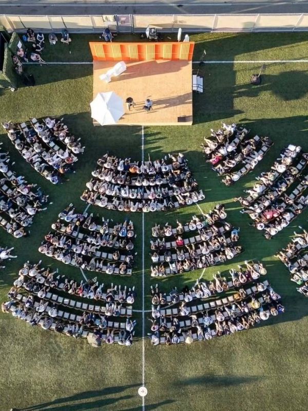 Overhead view of a large outdoor audience sitting in semicircles facing a stage on grass.