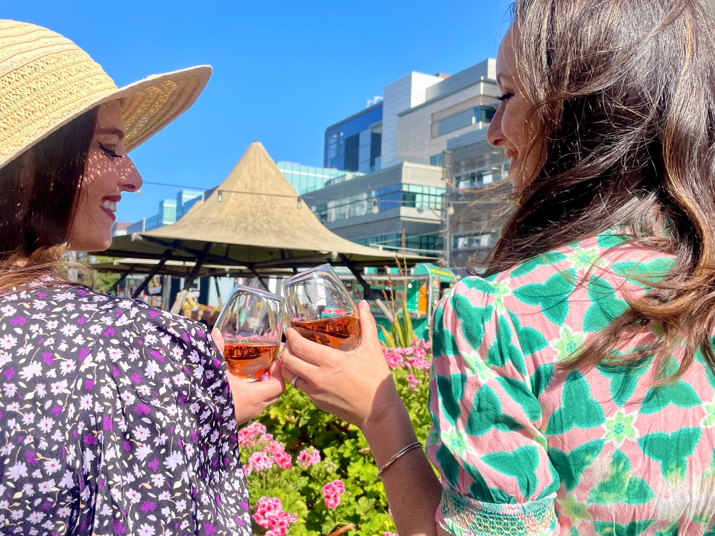 Two women toasting with drinks outdoors, surrounded by flowers and buildings under a clear blue sky.