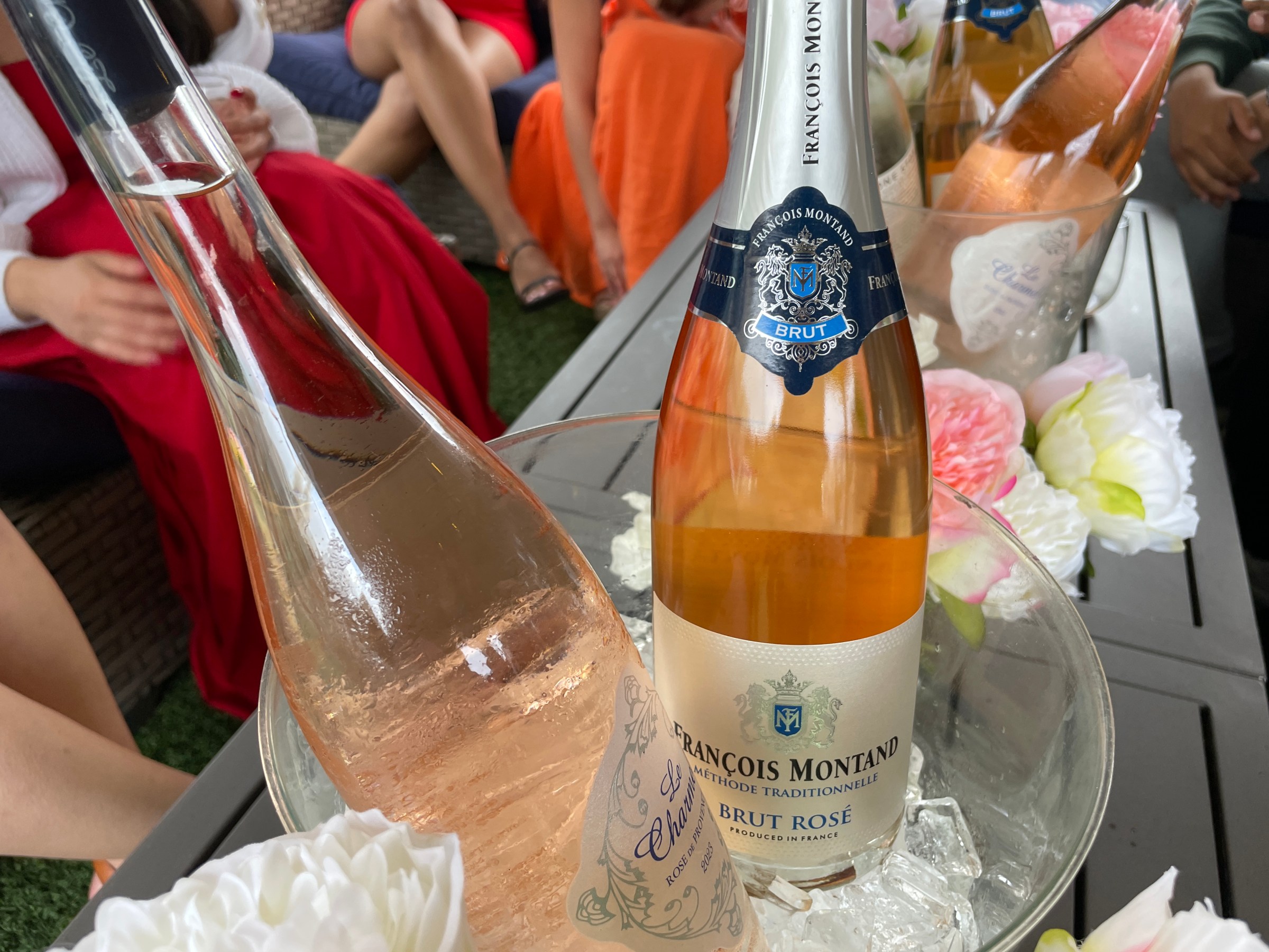 Bottles of Brut Rosé in ice bucket on table with flowers, people in colorful attire in background.