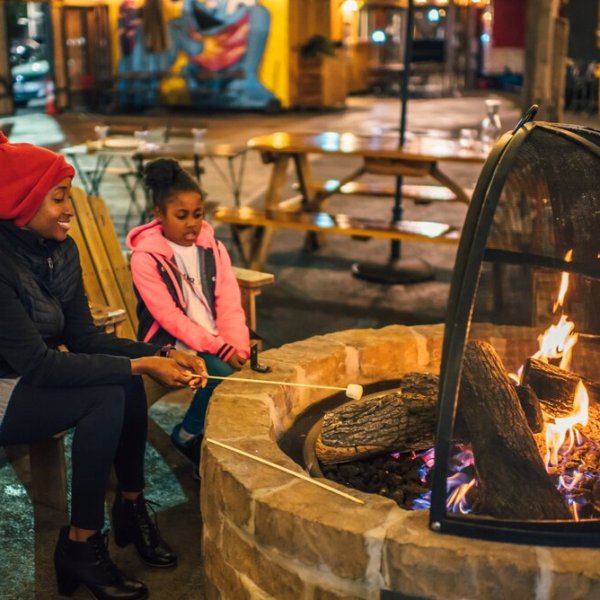 Two people roasting marshmallows over an outdoor fire pit with seating around.
