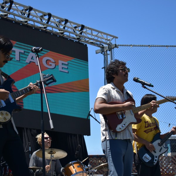 Band performing on outdoor stage with guitars and drums under a clear blue sky.