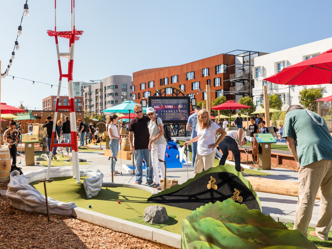 People playing miniature golf on a sunny day with buildings and trees in the background.